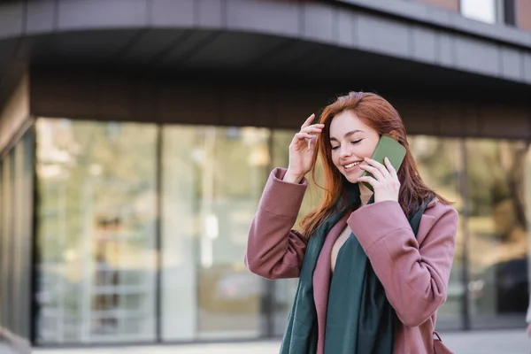 Щаслива руда жінка в шарфі і пальто розмовляє на мобільному телефоні зовні — стокове фото
