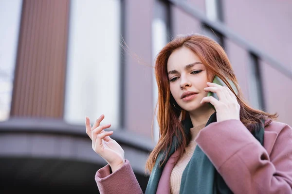Low angle view of young redhead woman in scarf and coat talking on smartphone outside — Stock Photo