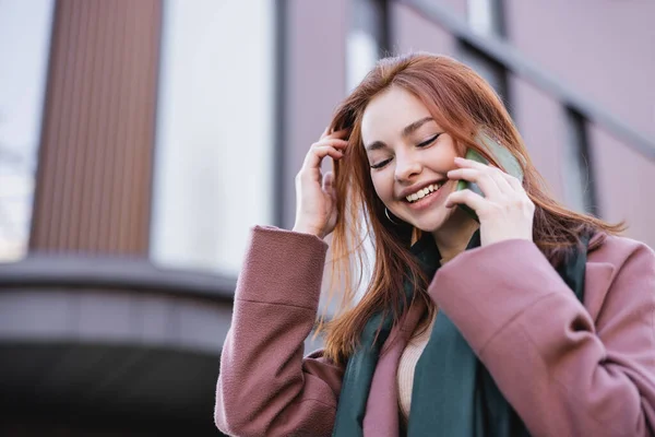 Vue à angle bas de femme rousse gaie en écharpe et manteau parlant sur smartphone à l'extérieur — Photo de stock