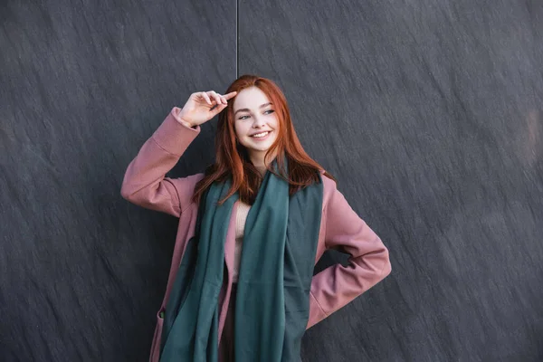 Happy redhead woman in scarf and coat smiling near grey marble wall — Stock Photo