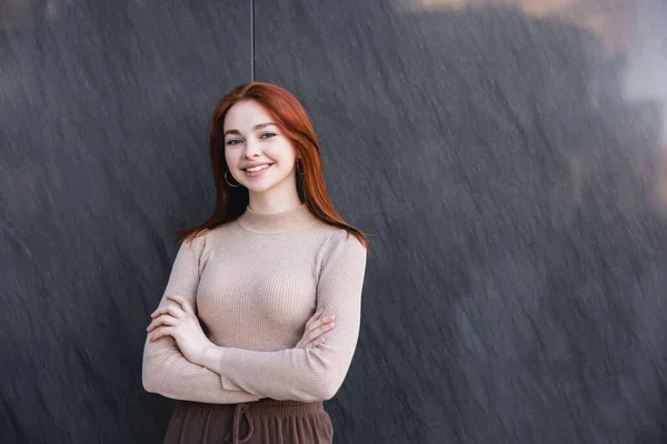 Happy redhead woman in beige turtleneck standing with crossed arms near grey marble wall - foto de stock