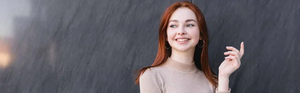 Joyful redhead woman in beige turtleneck gesturing near grey marble wall, banner — стоковое фото