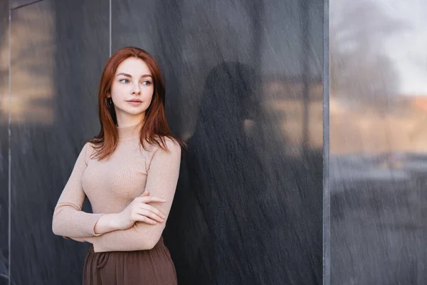 Young redhead woman in beige turtleneck standing with crossed arms near grey marble wall — стоковое фото