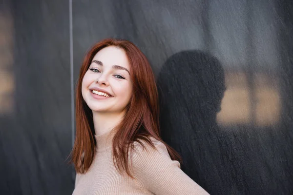 Happy redhead woman in beige turtleneck smiling near grey marble wall — стоковое фото