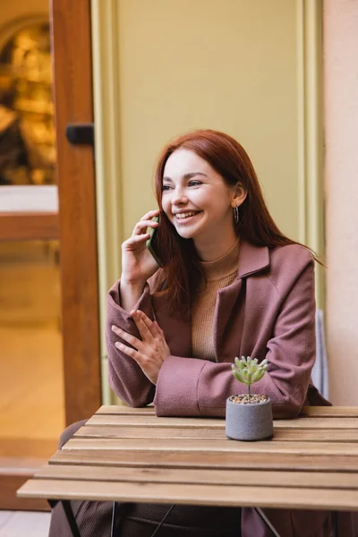 Mujer feliz con el pelo rojo hablando en el teléfono inteligente en la terraza de verano - foto de stock