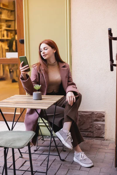 Full length of young woman with red hair using smartphone on summer terrace — Stock Photo