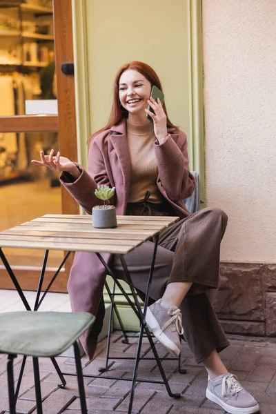 Longitud completa de la mujer feliz con el pelo rojo que tiene llamada telefónica en la terraza de verano - foto de stock