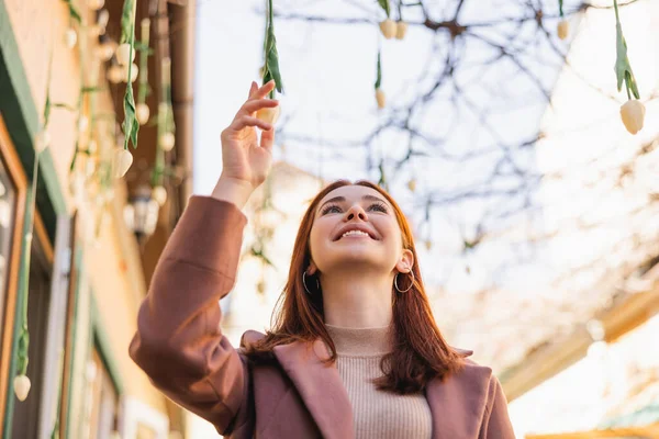 Low angle view of happy redhead woman in coat looking at hanging tulips outside — Stock Photo
