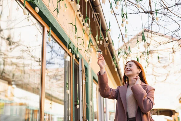 Low angle view of pleased redhead woman in coat smiling near hanging tulips outside — Stock Photo