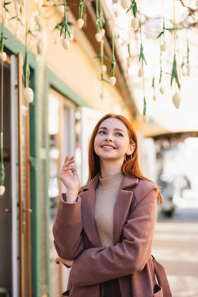 Dreamy redhead woman in coat smiling near hanging tulips outside — Stock Photo