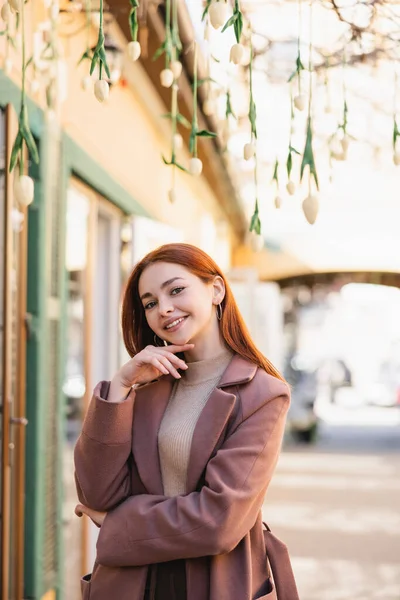 Happy young woman in coat smiling near hanging flowers outside — Stock Photo