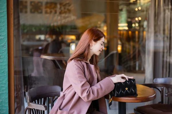 Young and happy woman with red hair looking at handbag in cafe terrace — Photo de stock