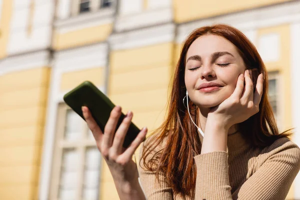 Cheerful woman listening music in wired earphones and holding smartphone — Stock Photo