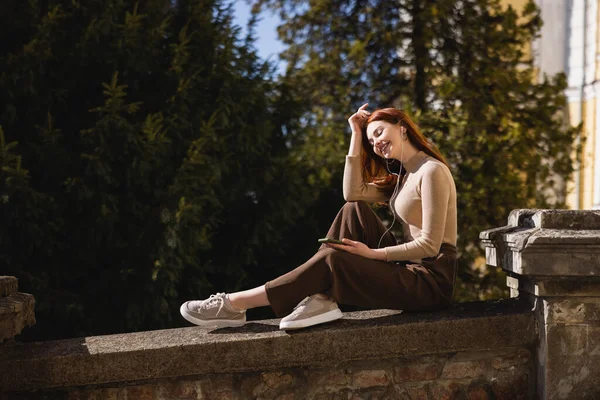 Cheerful woman listening music in wired earphones and holding smartphone while sitting outside — Photo de stock