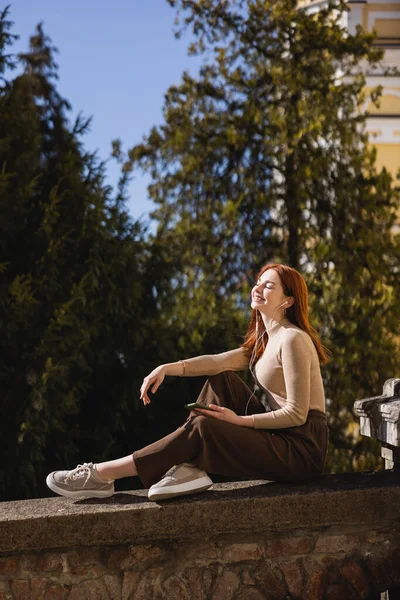 Pleased woman listening music in wired earphones and holding smartphone while sitting outside — стоковое фото