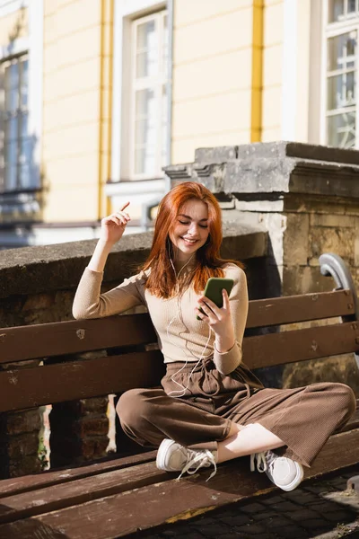 Pleased woman listening music in wired earphones and holding smartphone while sitting on bench — стоковое фото