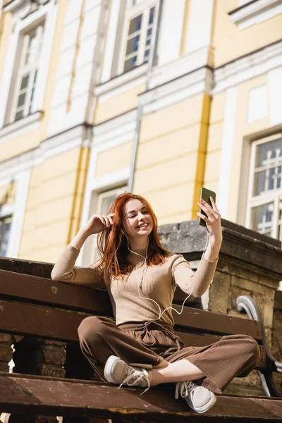 Pleased woman listening music in wired earphones taking selfie while sitting on bench - foto de stock