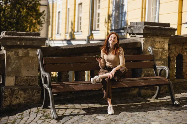 Pleine longueur de femme heureuse écoutant de la musique dans des écouteurs filaires et tenant smartphone tout en étant assis sur le banc — Photo de stock