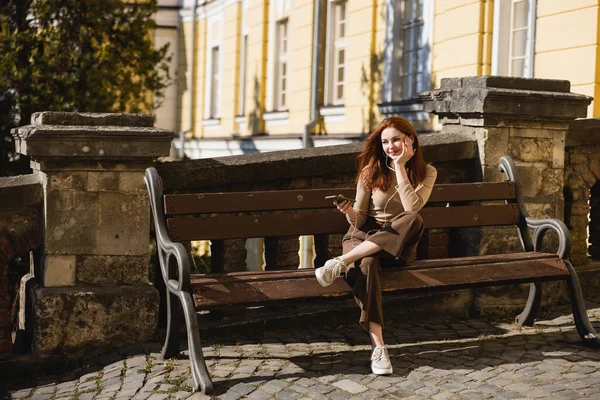 Pleine longueur de femme heureuse écoutant de la musique dans des écouteurs filaires et tenant smartphone tout en étant assis sur le banc — Photo de stock