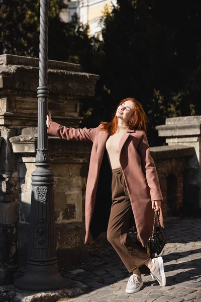 Sonne satt auf dem Gesicht einer glücklichen jungen Frau im Mantel mit Handtasche — Stockfoto