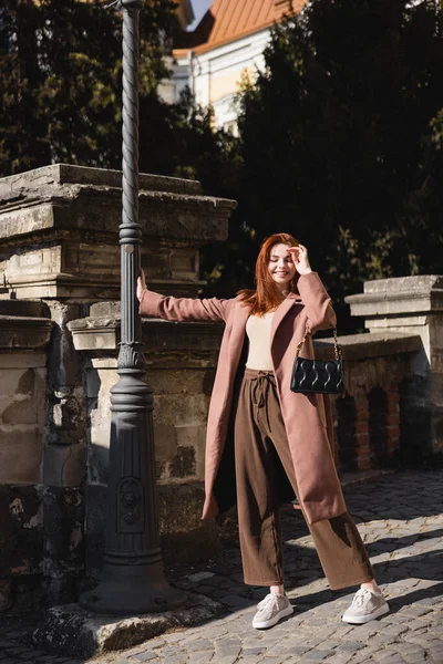Полная длина солнца на лице веселой молодой женщины в пальто держа сумочку — стоковое фото