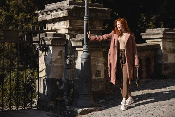 Full length of sunshine on face of happy young woman in coat holding handbag on street — Stock Photo