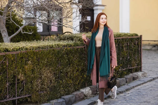 Longitud completa de la mujer joven en abrigo y bufanda sosteniendo el bolso y caminando en la calle - foto de stock