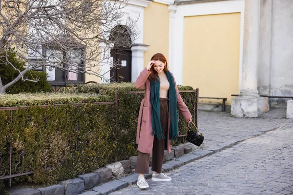 Повна довжина веселої молодої жінки в пальто і шарфі, тримаючи сумочку і гуляючи по вулиці — стокове фото
