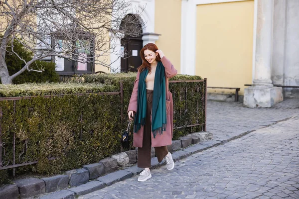 Full length of positive young woman in coat and scarf holding handbag and walking on street — стоковое фото