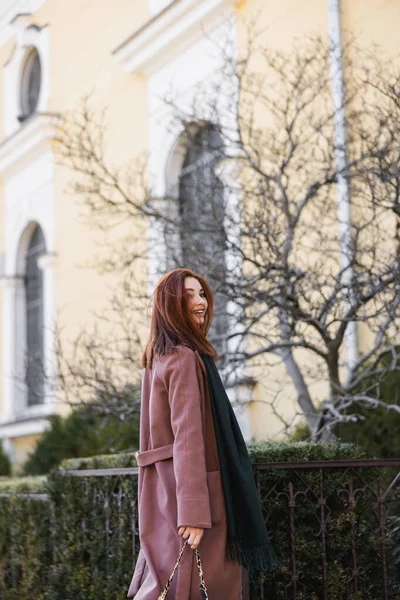 Mulher satisfeita em casaco e cachecol segurando bolsa e de pé na rua europeia — Fotografia de Stock