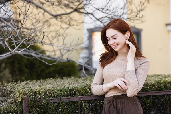 Happy and redhead woman adjusting wireless earphone outside — стоковое фото
