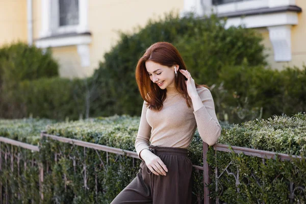 Young pleased woman in wireless earphone adjusting red hair outside — стоковое фото