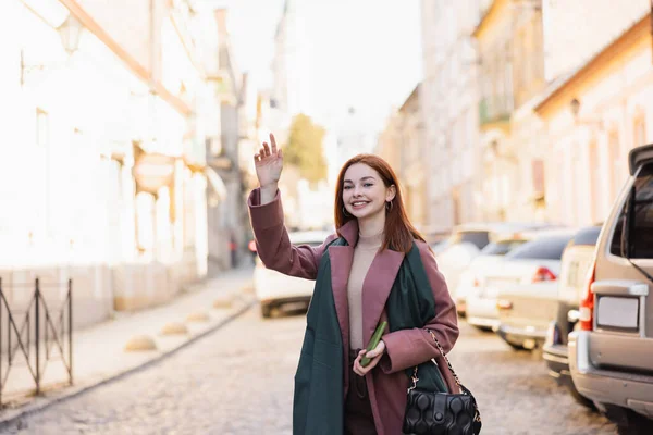 Cheerful redhead woman in coat calling taxi on european street - foto de stock