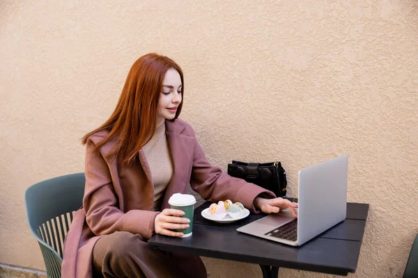 Joven pelirroja freelancer utilizando el ordenador portátil y la celebración de café para ir cerca de plato con macarrones - foto de stock