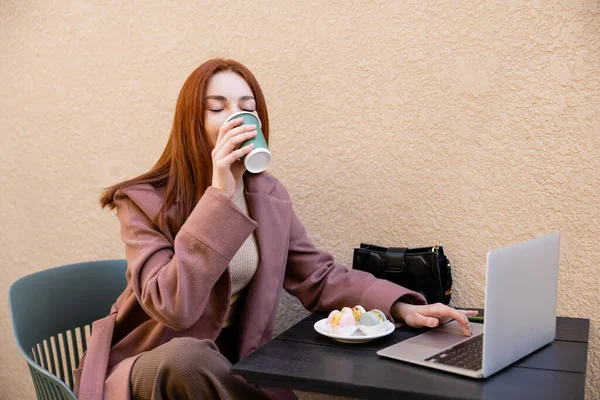 Young redhead freelancer drinking coffee to go near laptop and plate with macarons — Photo de stock