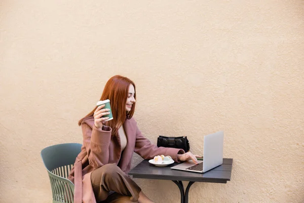 Joven freelancer feliz sosteniendo café para ir y mirando el ordenador portátil cerca de plato con macarrones - foto de stock