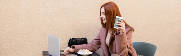 Happy freelancer holding coffee to go and using laptop near plate with macarons, banner — Stock Photo