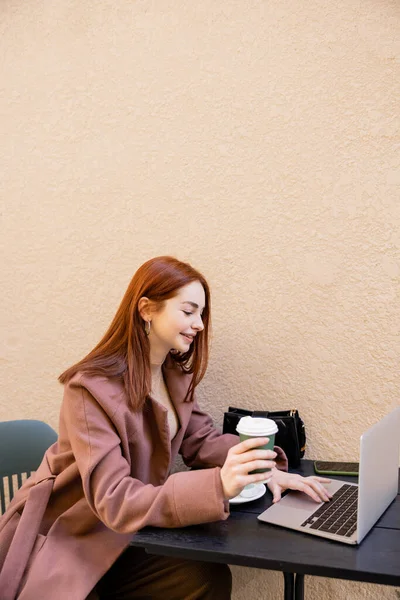 Cheerful woman holding coffee to go and using laptop on cafe terrace — Stock Photo