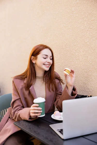 Femme gaie tenant du café pour aller et macaron près de l'ordinateur portable sur la terrasse du café — Photo de stock