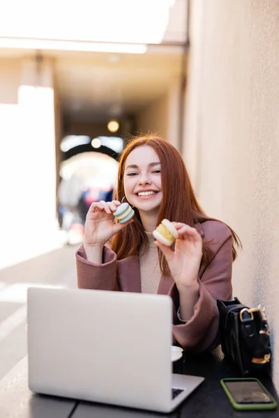 Femme gaie avec les cheveux roux tenant macarons doux près de l'ordinateur portable sur la terrasse du café — Photo de stock