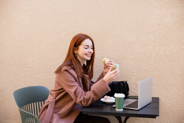 Happy young woman with red hair holding sweet macarons near laptop on cafe terrace — стоковое фото