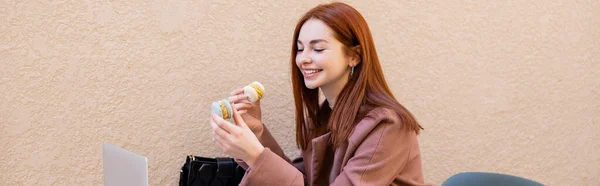 Heureuse jeune femme aux cheveux roux tenant des macarons doux près d'un ordinateur portable sur la terrasse du café, bannière — Photo de stock