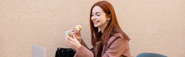 Feliz joven con el pelo rojo sosteniendo macarrones dulces cerca de la computadora portátil en la terraza de la cafetería, pancarta - foto de stock