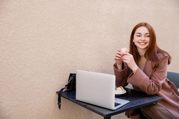 Joyful young woman with red hair holding sweet macaron near laptop on cafe terrace — Stock Photo