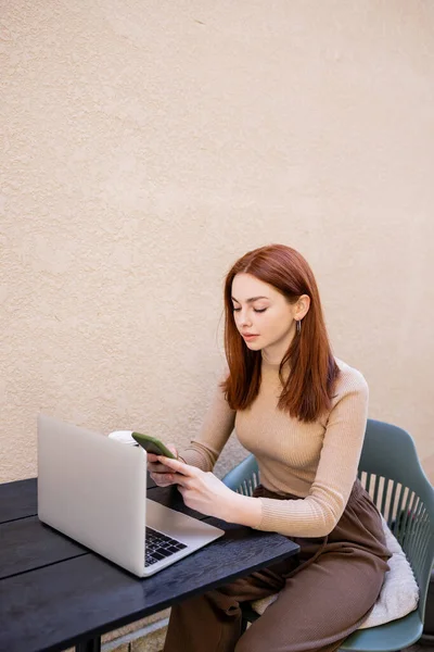 Junge rothaarige Frau benutzt Smartphone in der Nähe von Laptop auf Sommerterrasse — Stockfoto