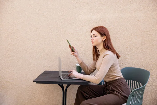 Young redhead freelancer in turtleneck using smartphone near laptop on summer terrace — Stock Photo