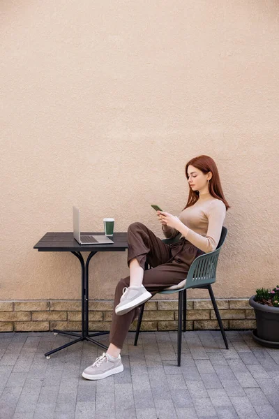 Full length of redhead woman in turtleneck using smartphone near laptop on summer terrace — стоковое фото