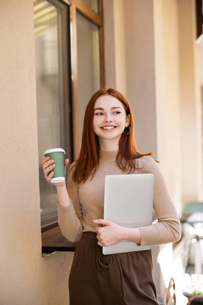 Young cheerful woman holding laptop and paper cup near building — Stock Photo