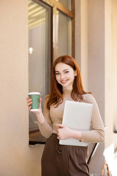 Smiling woman with red hair holding laptop and paper cup near building — Stock Photo