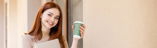 Щаслива молода жінка з рудим волоссям тримає ноутбук і паперову чашку біля будівлі, банер — стокове фото